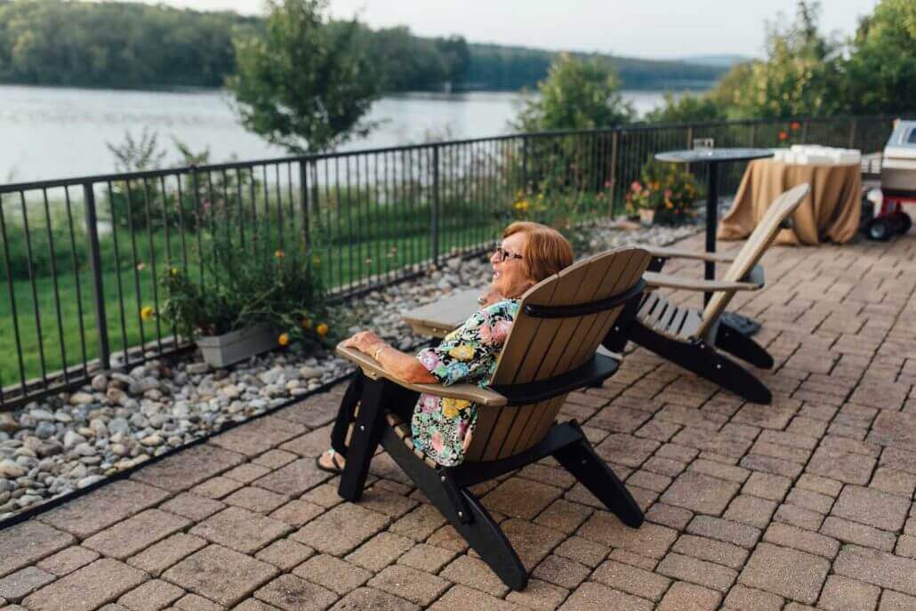 A woman sits in an adirondack chair on a patio at the Lake House Inn, overlooking a lake.