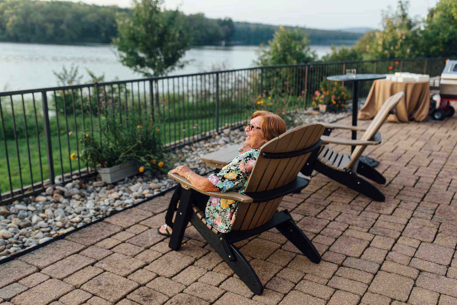 011-kate-eldar-lake-house-inn-rehearsal-dinner-philadelphia-wedding-photographer-alison-dunn-photography-photo A woman sits in an adirondack chair on a patio at the Lake House Inn, overlooking a lake.