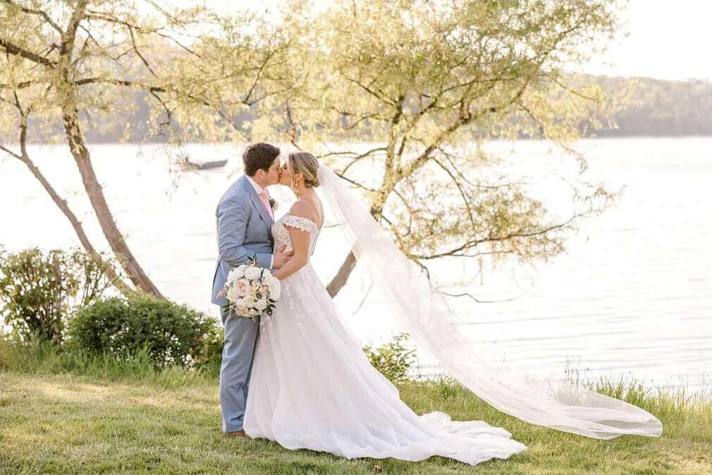 A bride and groom kiss in front of a lake during their wedding packages celebration.