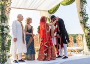 A couple in traditional attire exchange garlands at a Hindu wedding ceremony, with two people standing beside them under a decorated canopy, as the sacred water rituals unfold.