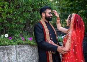 A couple in traditional attire stands outdoors, reminiscent of a Hindu wedding. The woman, wearing a red dress and veil, gently adjusts the man's glasses beside a stone wall with greenery.