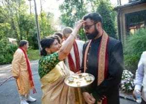 A woman marks a man's forehead during a traditional outdoor ceremony, reminiscent of Hindu wedding water rituals. In her other hand, she holds a tray with various items. Several people in traditional attire are present in the background.
